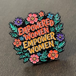 May include: A black enamel pin with the words "EMPOWERED WOMEN EMPOWER WOMEN" in orange and red lettering. The pin is surrounded by a floral wreath with pink, purple, and blue flowers and green leaves.