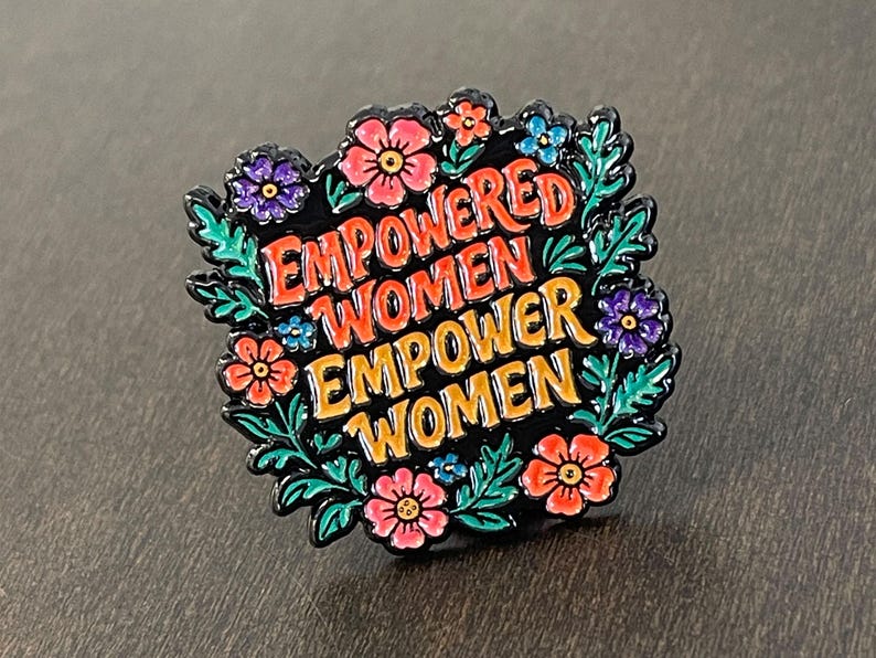 May include: A black enamel pin with the words "EMPOWERED WOMEN EMPOWER WOMEN" in orange and red lettering. The pin is surrounded by a floral wreath with pink, purple, and blue flowers and green leaves.