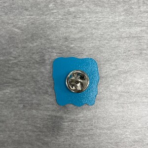K&ouml;nnte beinhalten: Ein t&uuml;rkisfarbener Emaille-Anstecker mit einem silberfarbenen Metall-Pin-R&uuml;cken. Der Anstecker hat eine einzigartige, unregelm&auml;&szlig;ige Form, und der Metall-Pin-R&uuml;cken ist rund mit einem Verschluss.