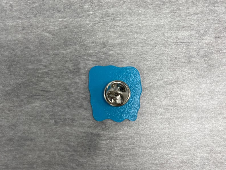 K&ouml;nnte beinhalten: Ein t&uuml;rkisfarbener Emaille-Anstecker mit einem silberfarbenen Metall-Pin-R&uuml;cken. Der Anstecker hat eine einzigartige, unregelm&auml;&szlig;ige Form, und der Metall-Pin-R&uuml;cken ist rund mit einem Verschluss.