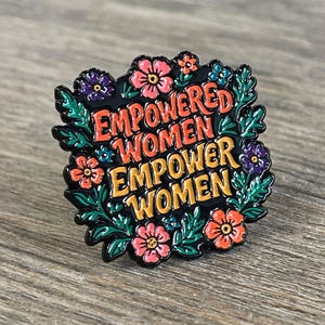 May include: A black enamel pin with the words "EMPOWERED WOMEN EMPOWER WOMEN" in orange and yellow lettering. The pin is surrounded by a floral design with pink, purple, and blue flowers and green leaves.