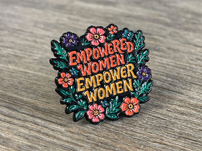 May include: A black enamel pin with the words "EMPOWERED WOMEN EMPOWER WOMEN" in orange and yellow lettering. The pin is surrounded by a floral design with pink, purple, and blue flowers and green leaves.