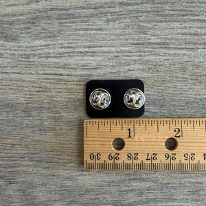 May include: Two silver-colored lapel pins with butterfly clasps are displayed on a black backing. A wooden ruler with inch markings is placed next to the pins, showing measurements.