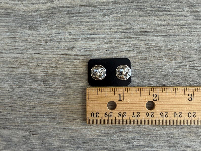 May include: Two silver-colored lapel pins with butterfly clasps are displayed on a black backing. A wooden ruler with inch markings is placed next to the pins, showing measurements.