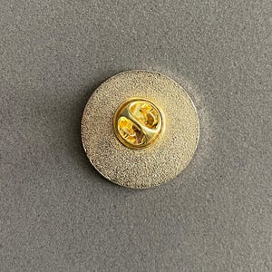 May include: A gold-colored round pin with a textured surface and a gold-colored pin back. The pin is circular and has a shiny, metallic appearance. The pin back has a butterfly clasp.