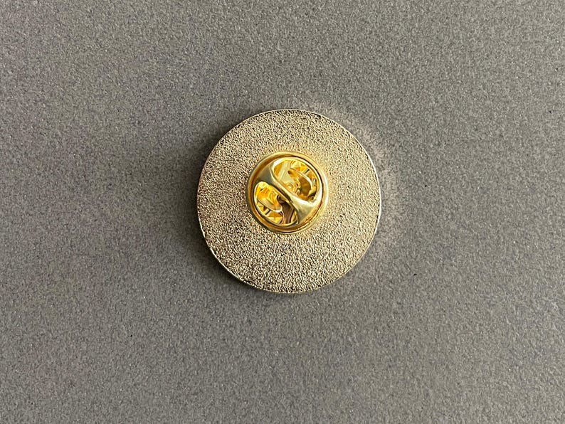 May include: A gold-colored round pin with a textured surface and a gold-colored pin back. The pin is circular and has a shiny, metallic appearance. The pin back has a butterfly clasp.