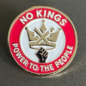 May include: Round pin with a gold border, red outer ring, and white center. The pin features a gold crown, a black fist, and the words "NO KINGS POWER TO THE PEOPLE" in white.