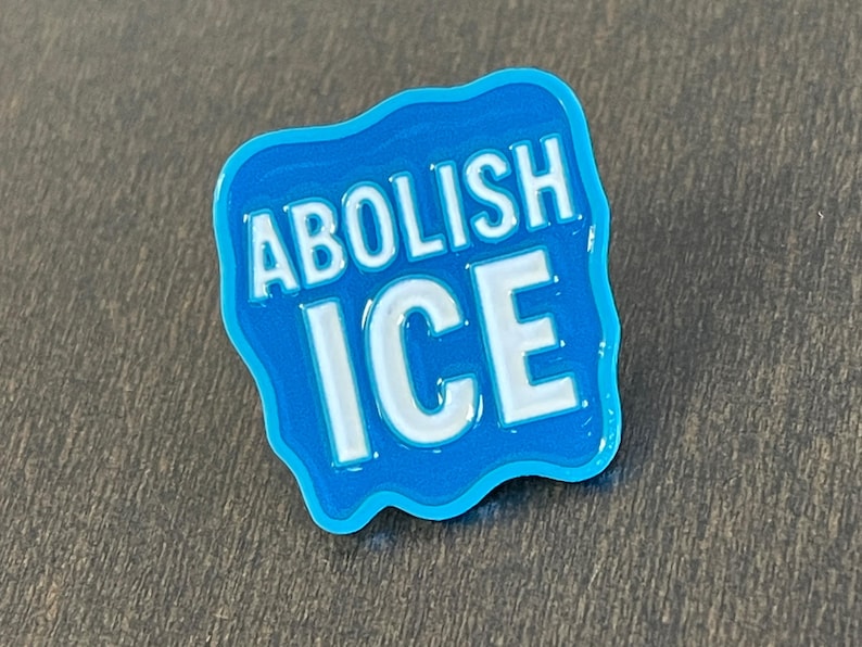 K&ouml;nnte beinhalten: Ein blauer Emaille-Pin mit den Worten "ABOLISH ICE" in wei&szlig;en Blockbuchstaben. Der Pin hat einen hellblauen Rand und eine wellenf&ouml;rmige Form und ist wahrscheinlich ein politisches Statement oder ein Protestartikel.