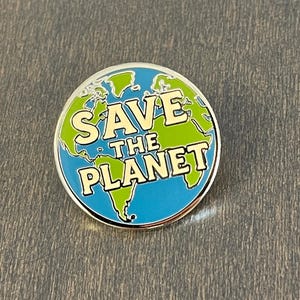 以下が含まれることがあります： 銀色の縁取りが付いた丸いエナメルピン。青と緑の地球のデザインに、白いブロック体で「SAVE THE PLANET」の文字が描かれています。ピンは灰色の背景に配置されています。