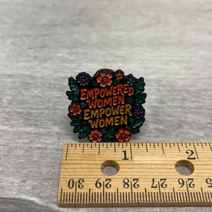 May include: A colorful enamel pin with the text "EMPOWERED WOMEN EMPOWER WOMEN" surrounded by flowers and leaves. The pin is black, red, green, and gold. A wooden ruler is next to the pin.