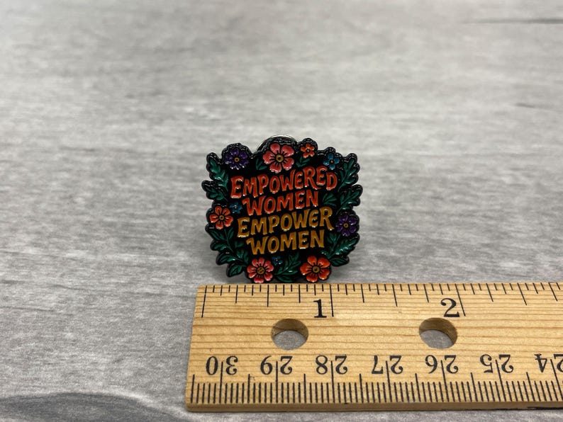 May include: A colorful enamel pin with the text "EMPOWERED WOMEN EMPOWER WOMEN" surrounded by flowers and leaves. The pin is black, red, green, and gold. A wooden ruler is next to the pin.