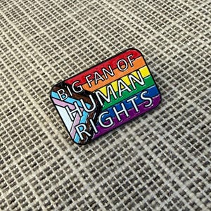 May include: A rectangular enamel pin with a rainbow design and the words "BIG FAN OF HUMAN RIGHTS." The pin includes the transgender pride flag colors and is outlined in black.