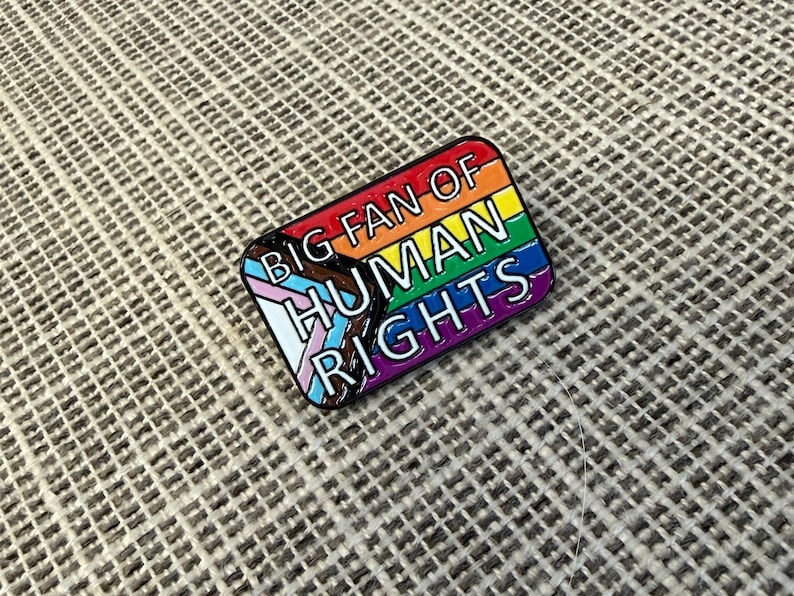 May include: A rectangular enamel pin with a rainbow design and the words "BIG FAN OF HUMAN RIGHTS." The pin includes the transgender pride flag colors and is outlined in black.