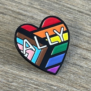 May include: A heart-shaped enamel pin with a black outline. The pin features the word "ALLY" in white letters over a rainbow design, including the colors of the LGBTQ+ pride flag and the transgender pride flag.