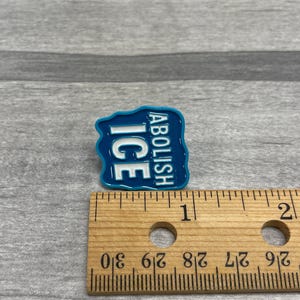 K&ouml;nnte beinhalten: Eine blaue Emaille-Anstecknadel mit den Worten "ABOLISH ICE" in wei&szlig;em Text. Die Anstecknadel ist etwa 2,5 cm breit und 2,5 cm hoch. Die Anstecknadel befindet sich auf einer grauen Oberfl&auml;che.