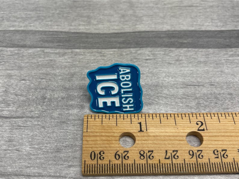 K&ouml;nnte beinhalten: Eine blaue Emaille-Anstecknadel mit den Worten "ABOLISH ICE" in wei&szlig;em Text. Die Anstecknadel ist etwa 2,5 cm breit und 2,5 cm hoch. Die Anstecknadel befindet sich auf einer grauen Oberfl&auml;che.