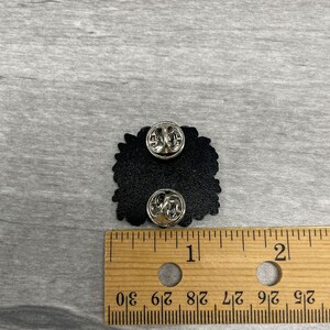 May include: Black enamel pin with two pin backs. The pin is approximately 1 inch wide. The pin is on a wooden ruler with inch markings. The pin is on a gray surface.