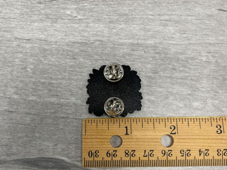 May include: Black enamel pin with two pin backs. The pin is approximately 1 inch wide. The pin is on a wooden ruler with inch markings. The pin is on a gray surface.