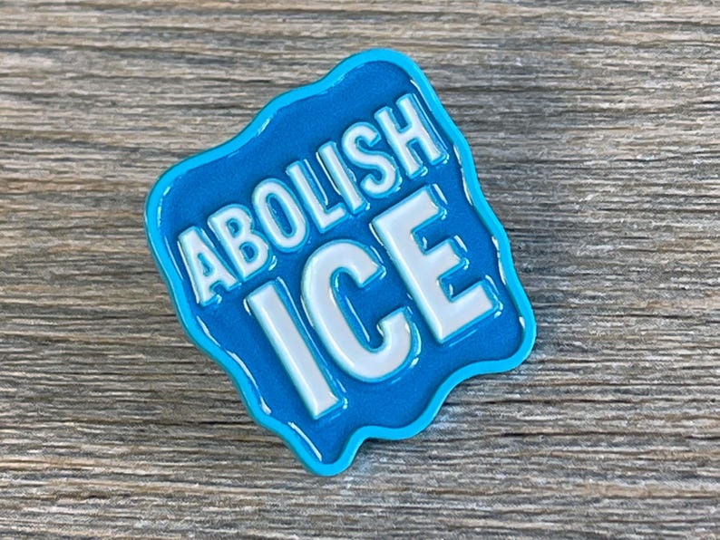 K&ouml;nnte beinhalten: Blauer Emaille-Pin mit den wei&szlig;en Worten "ABOLISH ICE". Der Pin hat einen welligen, unregelm&auml;&szlig;igen Rand und befindet sich vor einem Holzgrund.