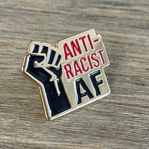 May include: A gold-colored enamel pin with a black fist and the words "ANTI-RACIST AF" in red and black. The pin is rectangular with a raised fist on the left and the text on the right. The pin is on a wooden surface.