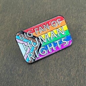 May include: A rectangular enamel pin with a rainbow flag design and the words "BIG FAN OF HUMAN RIGHTS" in white. The pin includes the Progress Pride flag and has a black border.