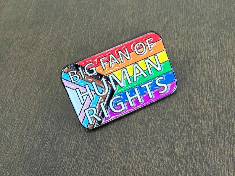 May include: A rectangular enamel pin with a rainbow flag design and the words "BIG FAN OF HUMAN RIGHTS" in white. The pin includes the Progress Pride flag and has a black border.