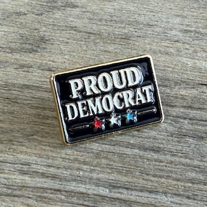 May include: A rectangular enamel pin with a black background and gold-tone metal border. The text PROUD DEMOCRAT is written in white capital letters. Below the text are three small stars colored red, white, and blue in a horizontal row.