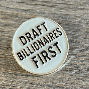 May include: A round, white enamel pin with the text "DRAFT BILLIONAIRES FIRST" in black, bold letters. The pin has a gold-colored border and is set against a wooden surface.