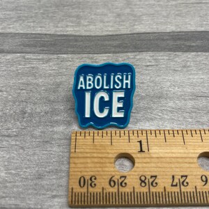 K&ouml;nnte beinhalten: Blauer Emaille-Anstecker mit dem wei&szlig;en Schriftzug &bdquo;ABOLISH ICE&ldquo;. Der Anstecker hat einen wellenf&ouml;rmigen Rand. Ein Holzlineal liegt neben dem Anstecker. Der Anstecker ist etwa 2,5 cm lang.