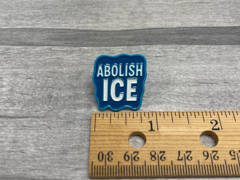 K&ouml;nnte beinhalten: Blauer Emaille-Anstecker mit dem wei&szlig;en Schriftzug &bdquo;ABOLISH ICE&ldquo;. Der Anstecker hat einen wellenf&ouml;rmigen Rand. Ein Holzlineal liegt neben dem Anstecker. Der Anstecker ist etwa 2,5 cm lang.