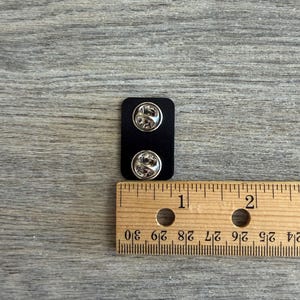 May include: A black rectangular pin back with two silver pin fasteners. A wooden ruler is placed next to the pin back, showing measurements in inches. The background is a gray wooden surface.