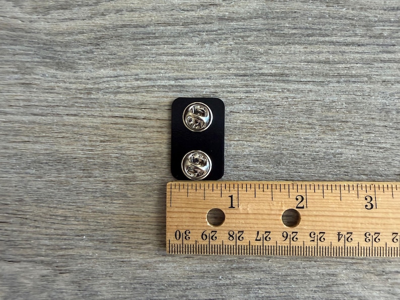 May include: A black rectangular pin back with two silver pin fasteners. A wooden ruler is placed next to the pin back, showing measurements in inches. The background is a gray wooden surface.