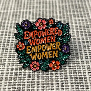 May include: A square enamel pin with the text "EMPOWERED WOMEN EMPOWER WOMEN" in orange and red lettering. The pin is surrounded by a wreath of green leaves and colorful flowers in shades of pink, purple, and blue.
