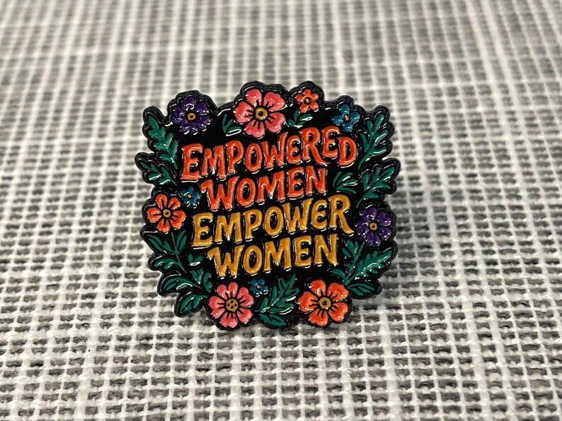 May include: A square enamel pin with the text "EMPOWERED WOMEN EMPOWER WOMEN" in orange and red lettering. The pin is surrounded by a wreath of green leaves and colorful flowers in shades of pink, purple, and blue.