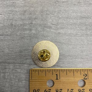 以下が含まれることがあります： ピンバック付きの金色の丸いピン。ピンの直径は約2.5cmです。木製の定規がスケールとしてピンの横に置かれています。