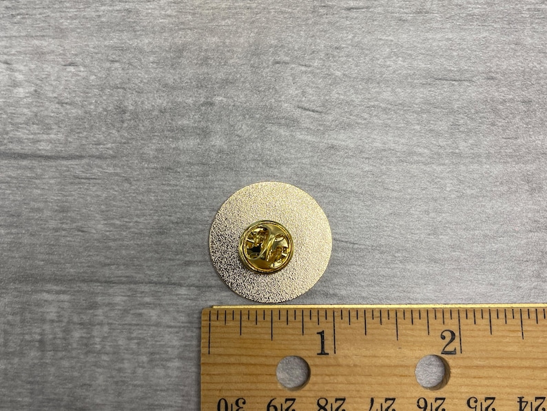 以下が含まれることがあります： ピンバック付きの金色の丸いピン。ピンの直径は約2.5cmです。木製の定規がスケールとしてピンの横に置かれています。
