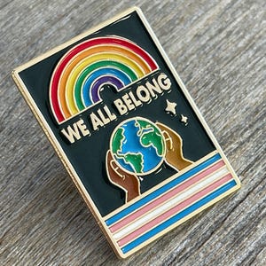 May include: Rectangular enamel pin with a gold border. The pin features a rainbow, the words "WE ALL BELONG", and a globe held by two hands. Below the globe is a transgender pride flag design.