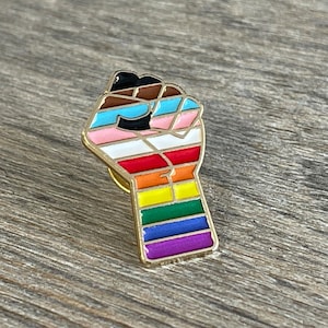 May include: A gold-colored enamel pin in the shape of a raised fist. The fist is segmented with various colors, including the colors of the LGBTQ+ pride flag, transgender flag, and other pride flags. The pin is set against a wooden background.