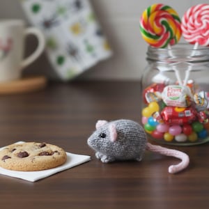 Könnte beinhalten: Eine handgestrickte graue Maus mit rosa Ohren und einem rosa Schwanz sitzt neben einem Schokoladen-Chip-Keks auf einer weißen Serviette. Ein Glas mit bunten Süßigkeiten und Lollis befindet sich im Hintergrund.