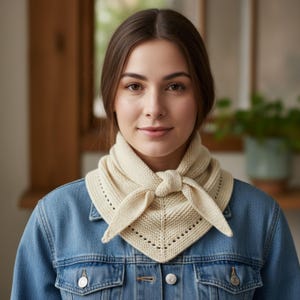May include: Cream-colored knitted scarf tied at the neck. The scarf has a triangular shape with a decorative border of small holes along the edges. The person is wearing a denim jacket.