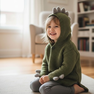 May include: A green knitted dinosaur hoodie with a hood featuring gray spikes and gray claws on the sleeves. The button-up sweater is worn by a child. The child is wearing gray pants and is sitting on a light-colored rug.