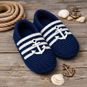 May include: A pair of navy blue knitted slippers with white stripes and a white anchor emblem. The nautical-themed slippers are displayed on a wooden surface, with seashells, rope, and decorative anchors.