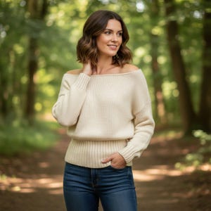 May include: Off-the-shoulder cream-colored sweater with ribbed detailing at the neckline, cuffs, and waist. The sweater is paired with blue jeans. The background is a blurred forest scene.