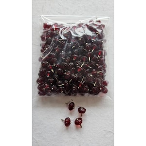 May include: A clear plastic bag filled with numerous dark red, flower-shaped beads. Four individual beads are scattered below the bag. Each bead has a small, silver-colored pin attached to the back.