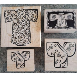 May include: Four wooden stamps with kimono designs. The stamps feature black ink illustrations of kimono robes. One stamp has a detailed pattern, while others have floral or bamboo motifs. The stamps are on a light-colored wooden base.
