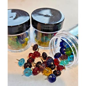 May include: A collection of colorful glass pumpkin-shaped push pins in various colors, including blue, red, yellow, and green. The pins are scattered around and inside clear plastic jars with black lids.