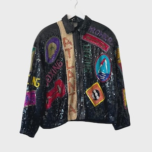 May include: Black sequined jacket with colorful patches and text. Patches include the words "Atlanta" and "Boxing", along with other graphics. The jacket has a zip-up front and a beige stripe down the center.