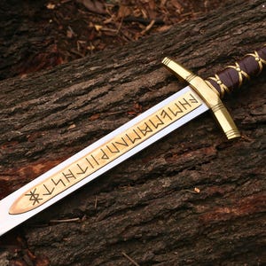 May include: A detailed image of a sword with a gold hilt and crossguard, a brown leather-wrapped handle, and a blade with engraved text. The sword rests on a textured, brown wooden surface.