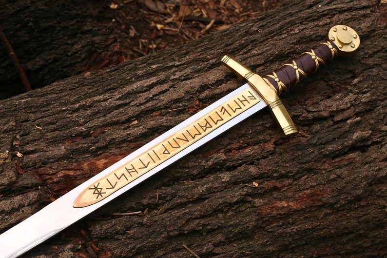 May include: A detailed image of a sword with a gold hilt and crossguard, a brown leather-wrapped handle, and a blade with engraved text. The sword rests on a textured, brown wooden surface.