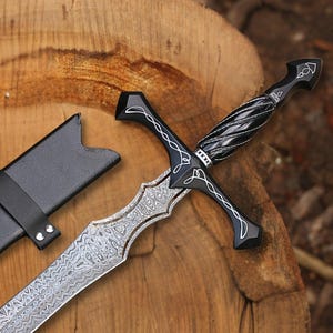Può includere: Una spada decorativa con una lama d'argento e un'impugnatura nera. L'impugnatura ha un design attorcigliato e dettagli bianchi. La spada è appoggiata su una superficie di legno accanto a un fodero nero.